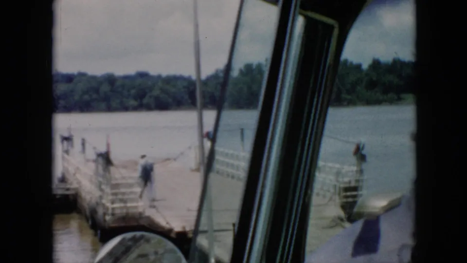 Inside-vehicle view of a 1956 Cuba ferry landing, with window frame, side mirror, worker, pier railings, and river beyond.