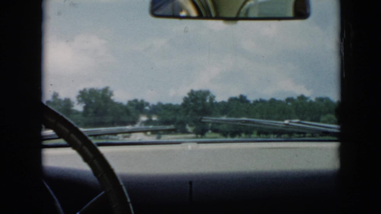 1956 Cuba home-movie frame from inside a car, looking past the steering wheel and dashboard toward a tree-lined road and sky.