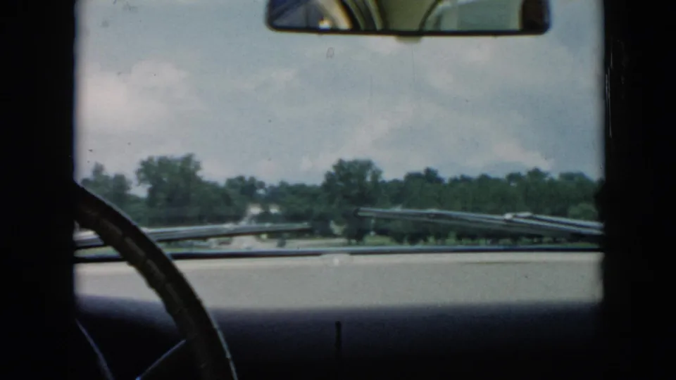 1956 Cuba home-movie frame from inside a car, looking past the steering wheel and dashboard toward a tree-lined road and sky.