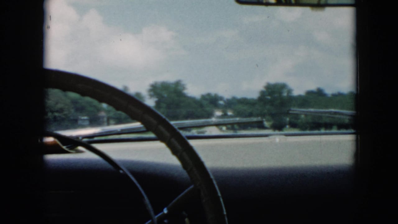 1956 Cuba home-movie view from inside a moving car, with the steering wheel silhouetted against roadside trees and sky.