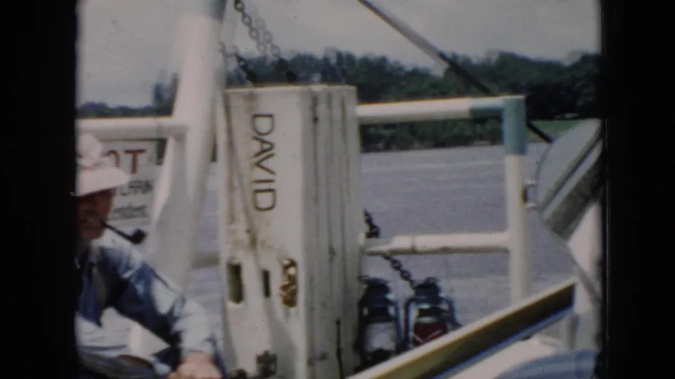 1956 Cuba home-movie frame of an adult man in a white hat seated aboard a boat near railings and a post marked DAVID.