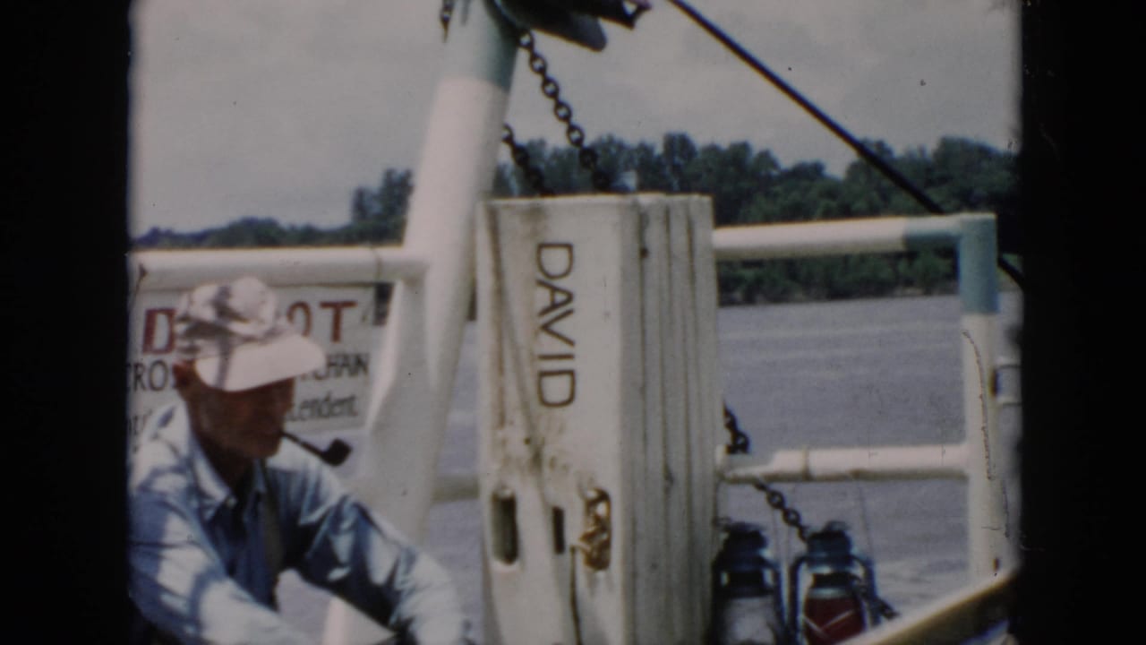 In 1956 Cuba home-movie footage, a man in a pale hat and blue shirt sits on a boat near water and a post marked DAVID.