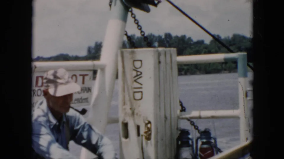 In 1956 Cuba home-movie footage, a man in a pale hat and blue shirt sits on a boat near water and a post marked DAVID.