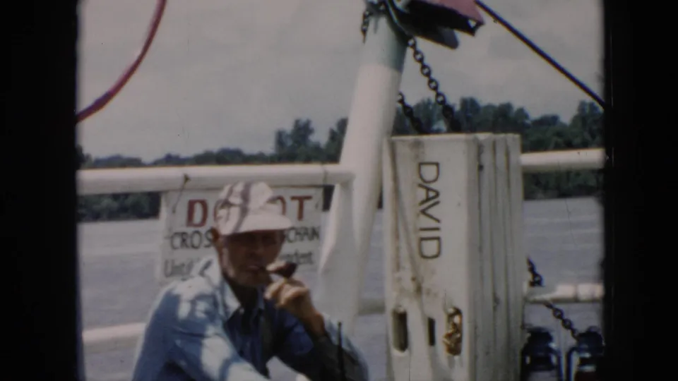 The man appears to smoke or hold a pipe on the boat, with white railings, the DAVID marking, water, and trees in the background.