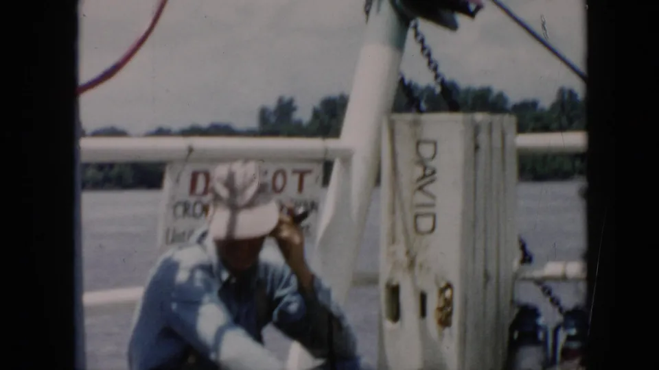 The man lowers his head and touches his hat on the boat, with the DAVID-marked post, railings, water, and shoreline behind him.