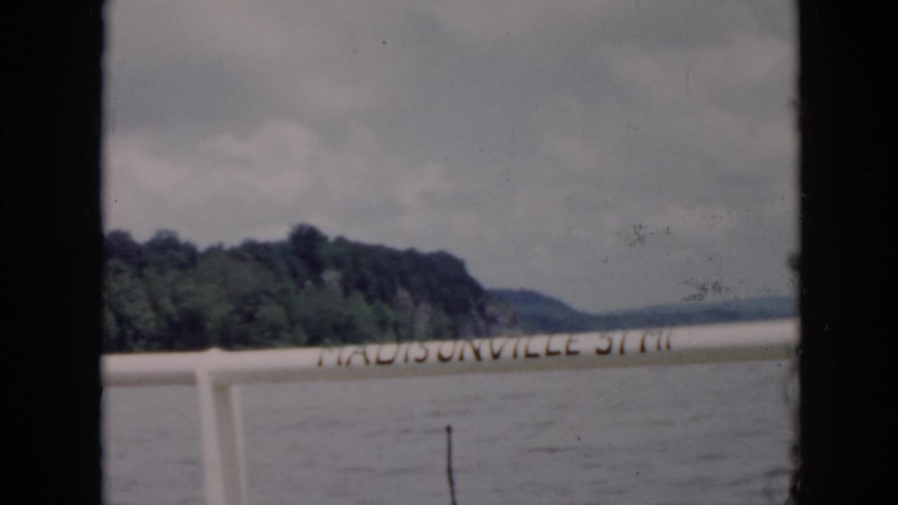 1956 8mm home-movie frame identified as Cuba, with a Madisonville-marked rail or sign against water and a tree-covered shore.
