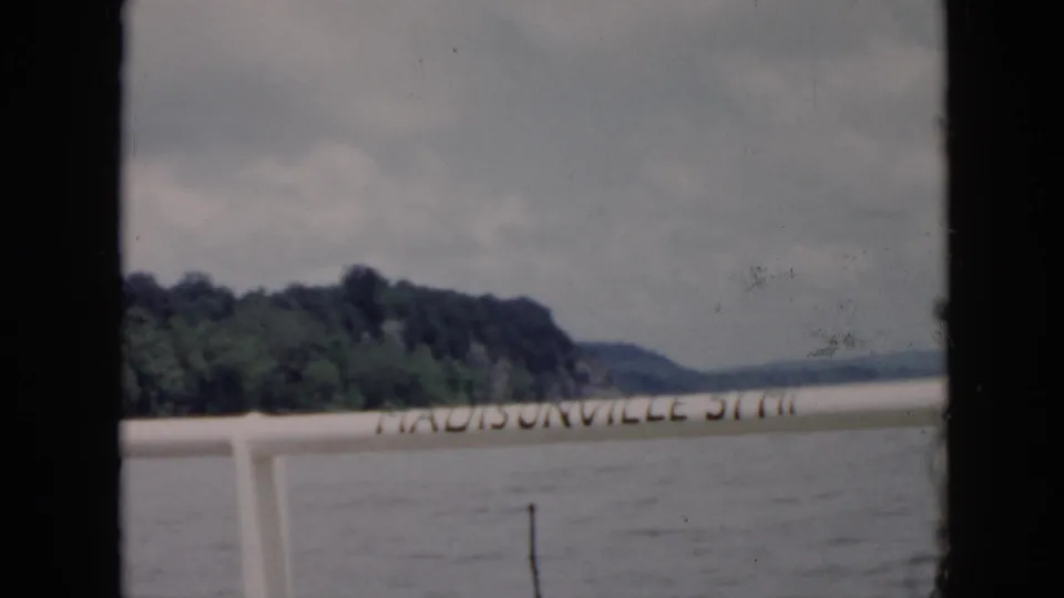 1956 8mm home-movie frame identified as Cuba, with a Madisonville-marked rail or sign against water and a tree-covered shore.