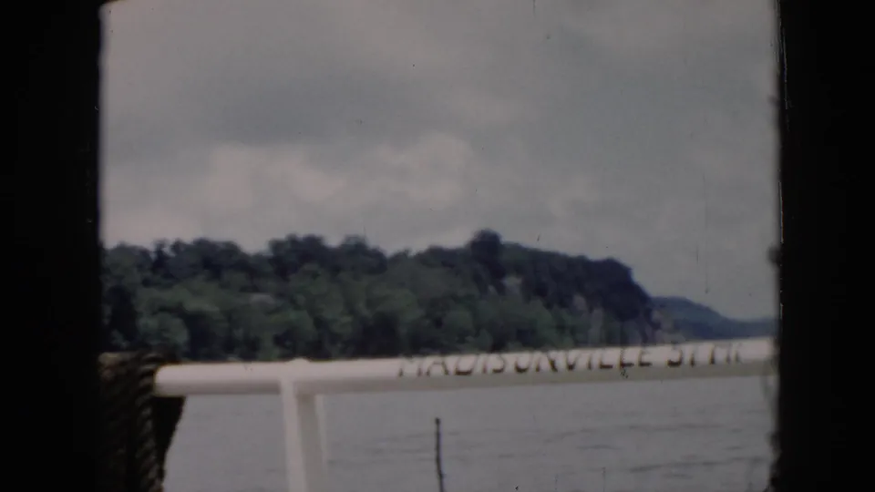 1956 8mm home-movie frame identified as Cuba, with boat railing, Madisonville lettering, water, and a wooded shoreline in soft focus.