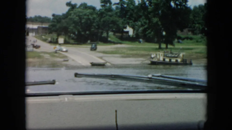 1956 Cuba shoreline view from a moving boat, with a landing road, parked cars, trees, and a small tug-like vessel.