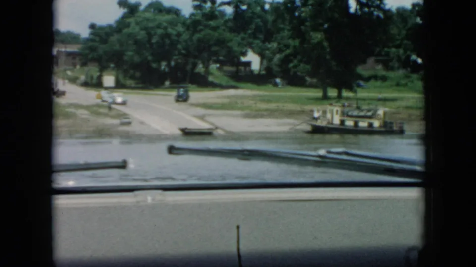 Late clip frame from a 1956 Cuba boat ride, showing the shore landing, vintage cars, green trees, and rails from the filming boat.