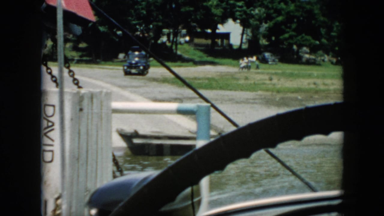 View through a vehicle toward a riverside road, dark car, pier, small boat, trees, and buildings.