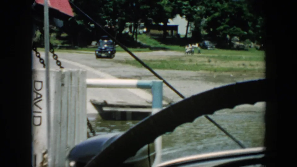 1956 Cuba home-movie frame looking past a steering wheel toward a riverside road with a dark car, pier, small boat, trees, and buildings.