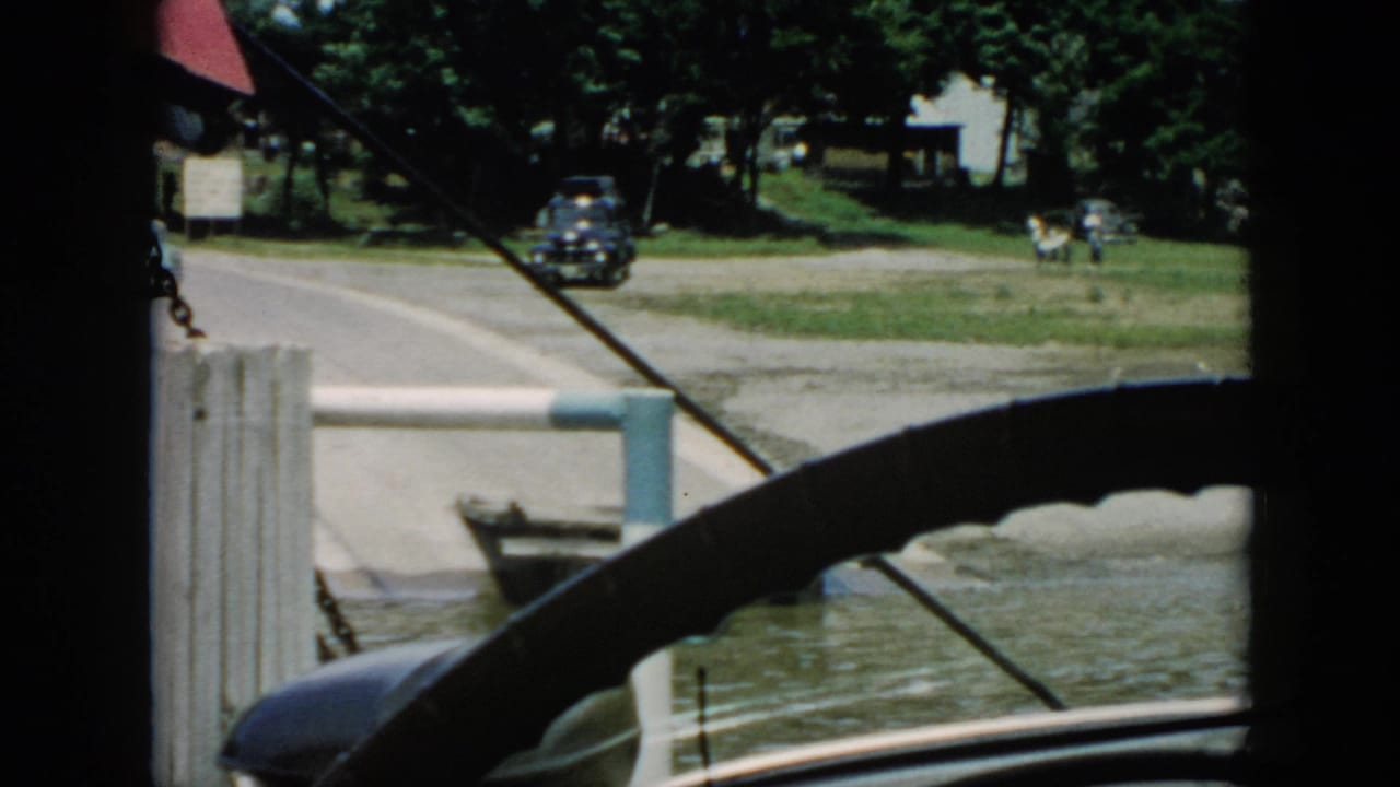 1956 Cuba travel frame seen from inside a vehicle, showing a dark car near a riverside road, dock structure, water, trees, and distant buildings.