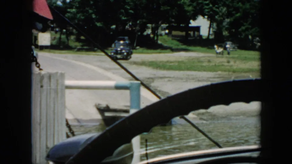 1956 Cuba travel frame seen from inside a vehicle, showing a dark car near a riverside road, dock structure, water, trees, and distant buildings.