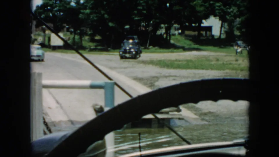 1956 Cuba frame from a moving vehicle, with a light car at the road edge, a dark car ahead, dock, river water, grassy bank, and trees.