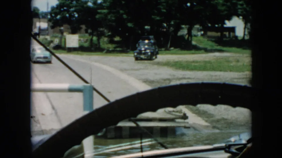 1956 Cuba home-movie view through a vehicle, showing a road beside water, a dark car, dock, grassy bank, trees, buildings, and another car passing.