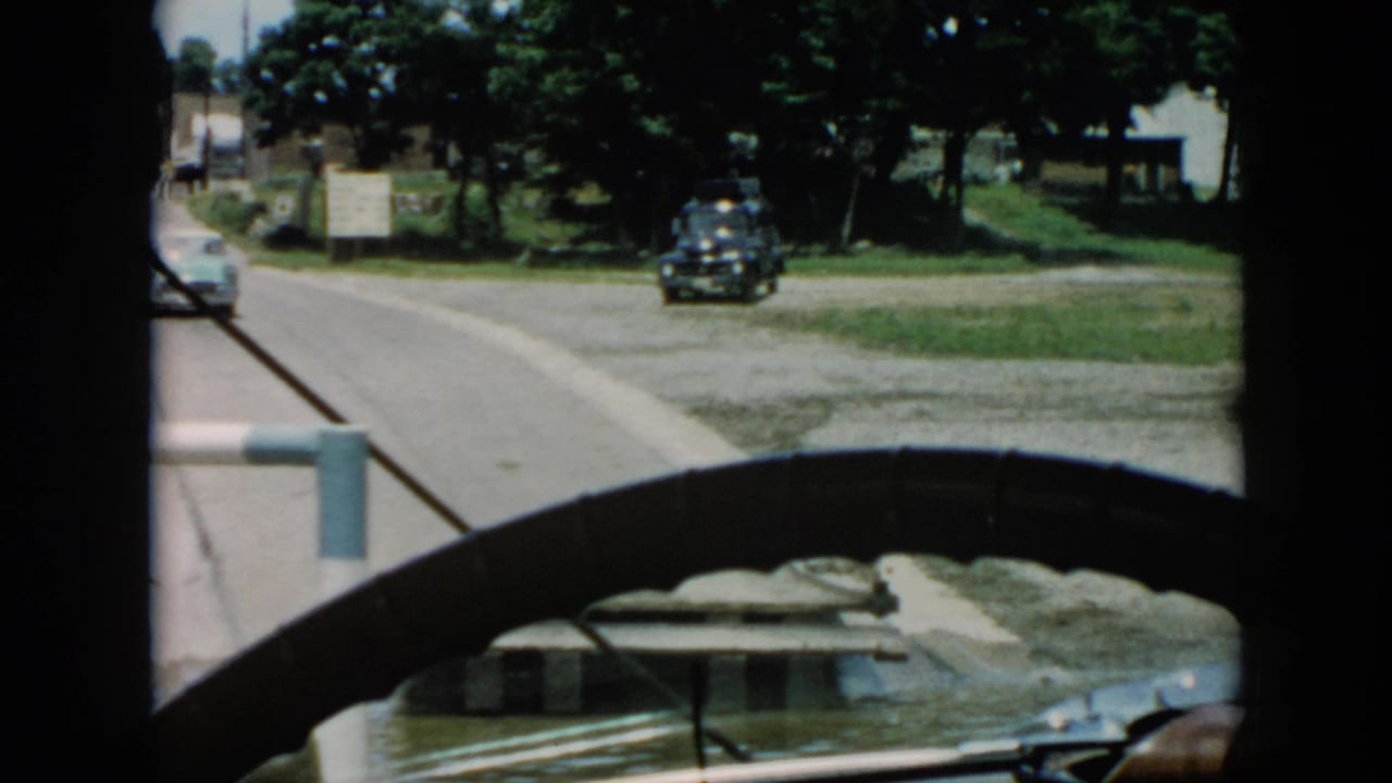1956 Cuba 8mm frame looking through a car windshield toward a road, a light-colored car ahead, trees, grass, signs, and a dark parked vehicle.