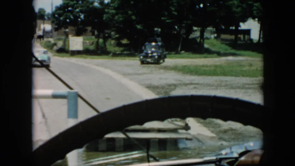 1956 Cuba 8mm frame looking through a car windshield toward a road, a light-colored car ahead, trees, grass, signs, and a dark parked vehicle.