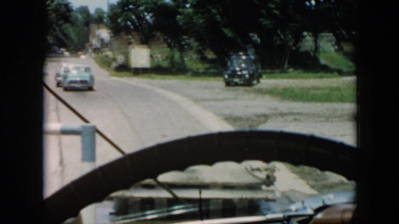 A light-colored car drives ahead on a road seen through a dark windshield frame.