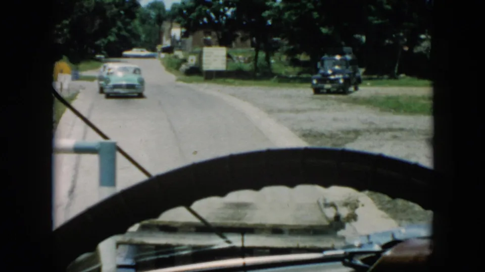 1956 Cuba 8mm frame showing a windshield view of a car ahead near crosswalk markings, with trees, grass, signs, and another dark vehicle visible.