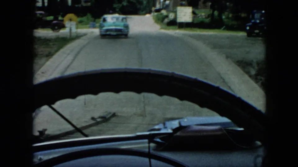 1956 Cuba home-movie frame with a teal car centered on a quiet road, seen through the windshield with dashboard edges and roadside trees visible.