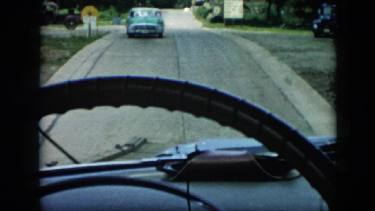1956 Cuba 8mm frame from inside a car, with the steering wheel and dashboard framing a quiet road and blue automobile ahead.