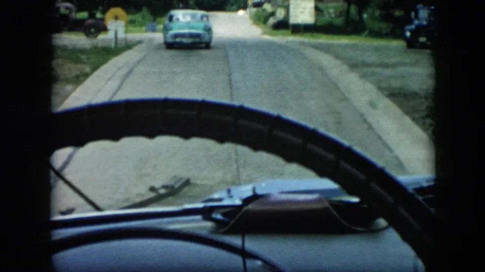 1956 Cuba 8mm frame from inside a car, with the steering wheel and dashboard framing a quiet road and blue automobile ahead.