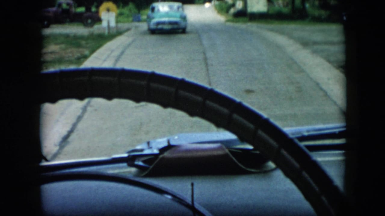 Steering wheel and dashboard frame a road scene with a blue car in the distance.