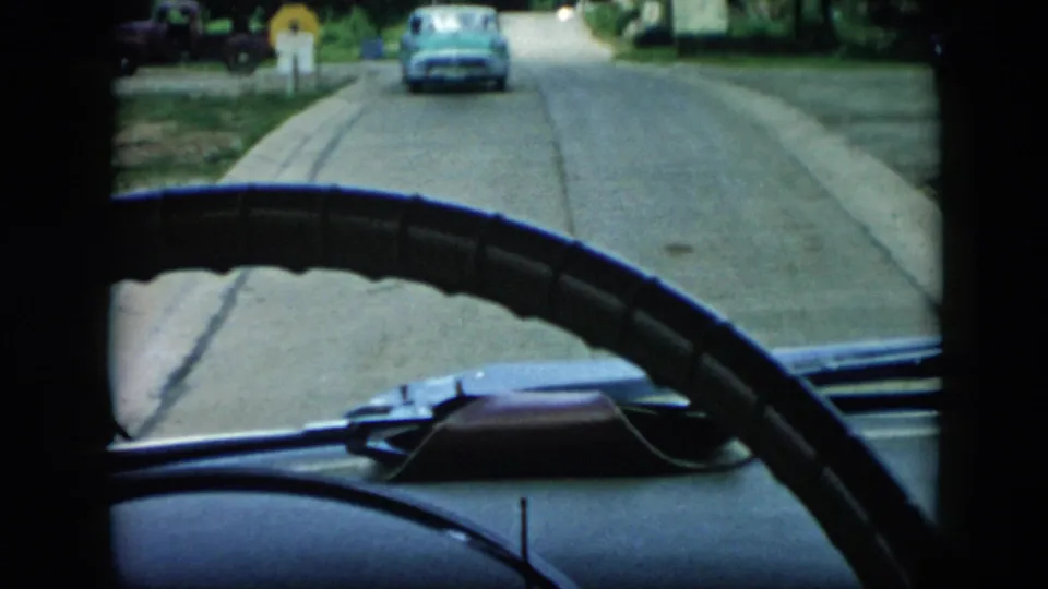 Car windshield view in 1956 Cuba, showing a classic steering wheel, dashboard edge, road, and blue mid-century car ahead.