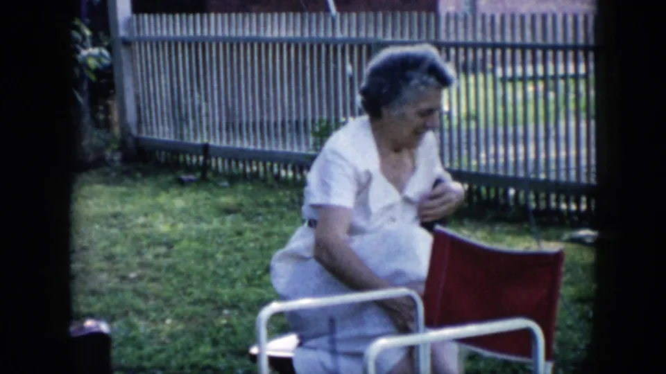 1956 Cuba home-movie frame showing a woman in a white dress seated outdoors on a red chair beside a fence and grass.