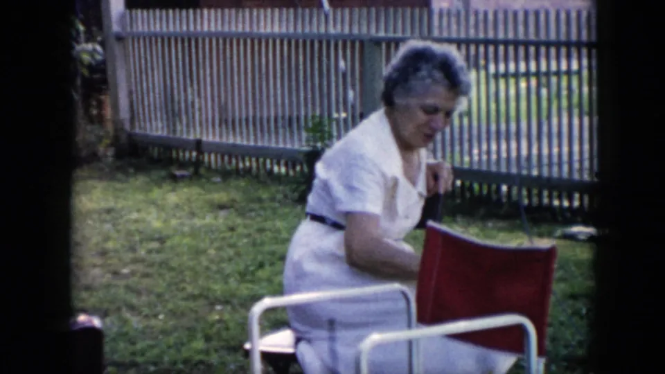 Home-movie frame from 1956 Cuba with a woman seated in a yard, looking down while sitting on a red folding chair near a fence.