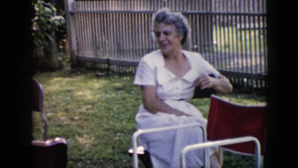 An older woman in a white dress smiles while seated in a red folding chair in the 1956 Cuba backyard scene.