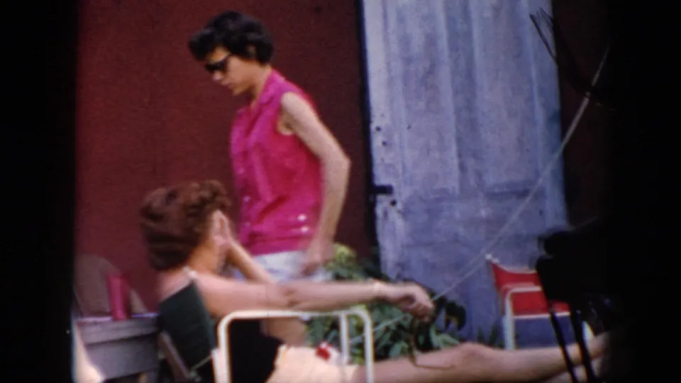 A younger woman in a bright pink sleeveless top walks past another seated woman near chairs in the backyard.