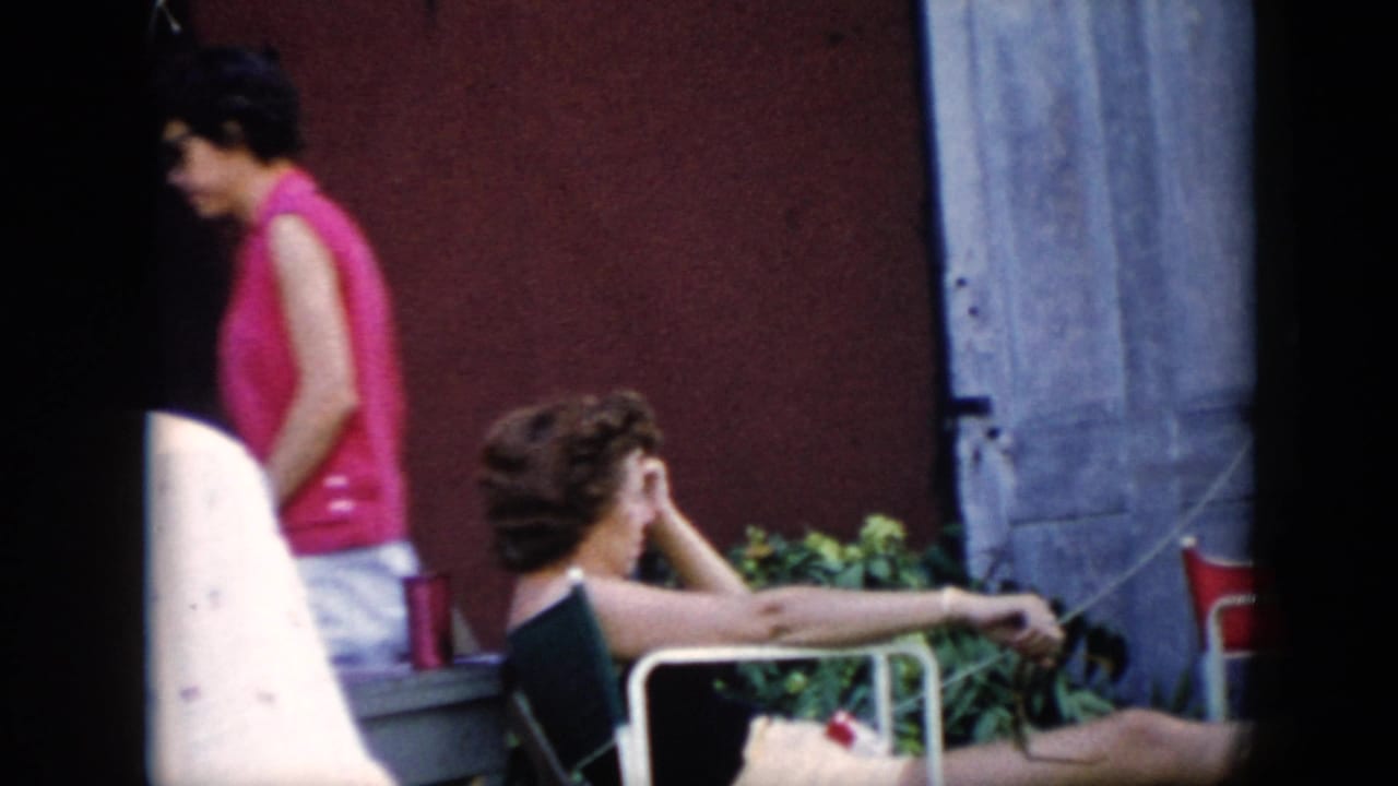 In Cuba home-movie footage from 1956, one woman in pink passes a reclining woman seated outdoors near a red wall and blue shutter.