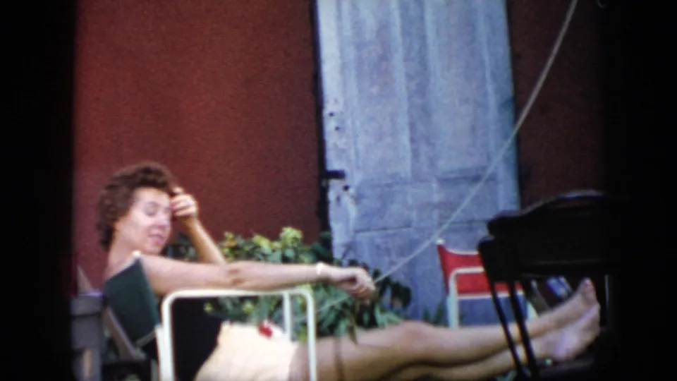 The 1956 Cuba home-movie frame shows a woman reclining in a backyard chair, partly shaded by the camera framing and nearby wall.