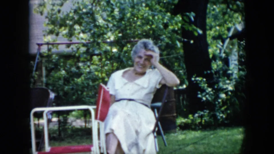 A woman in a white dress sits smiling toward the camera in a Cuba garden, with folding chairs, lawn, and dense foliage behind her.