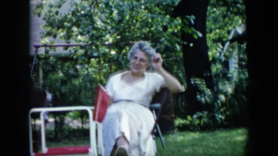 In 1956 Cuba home-movie footage, an older woman in a white dress sits in a green backyard and looks toward the camera.