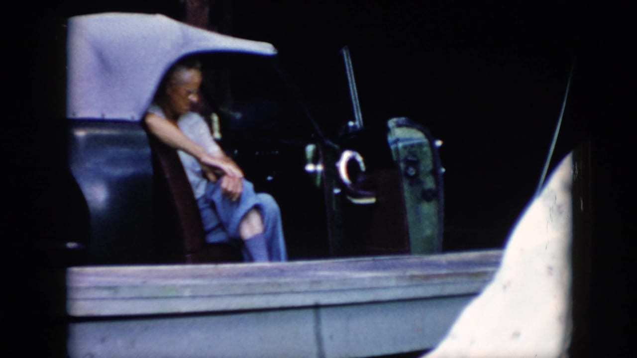 A man sits in a dark open car with a pale soft top, filmed from a home-movie perspective in the supplied Cuba 1956 clip.