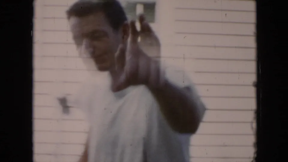 A younger man in a light shirt waves at the camera in the Cuba 1956 home-movie footage, with a pale wall behind him.