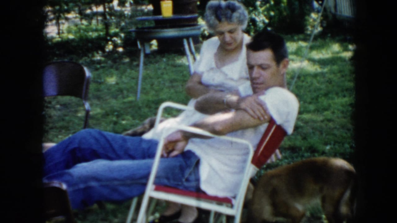 1956 Cuba home-movie frame of two adults relaxing in a shaded grassy backyard as a brown dog moves near the seated man.