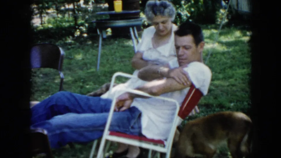 1956 Cuba home-movie frame of two adults relaxing in a shaded grassy backyard as a brown dog moves near the seated man.