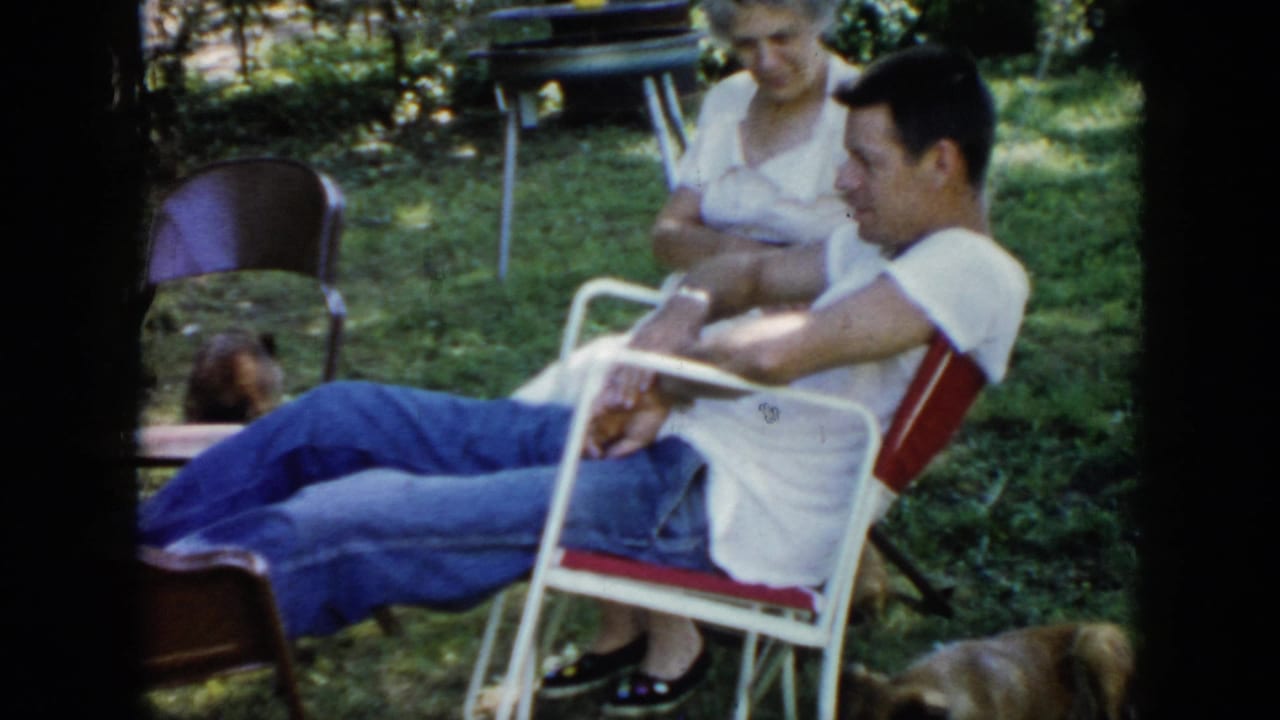 Man reclines in a folding chair with a woman behind him and a dog at the edge of the yard.