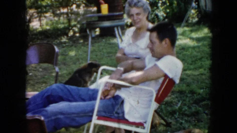 1956 Cuba home-movie frame of a brown dog near two adults seated outdoors among folding chairs and backyard shade.