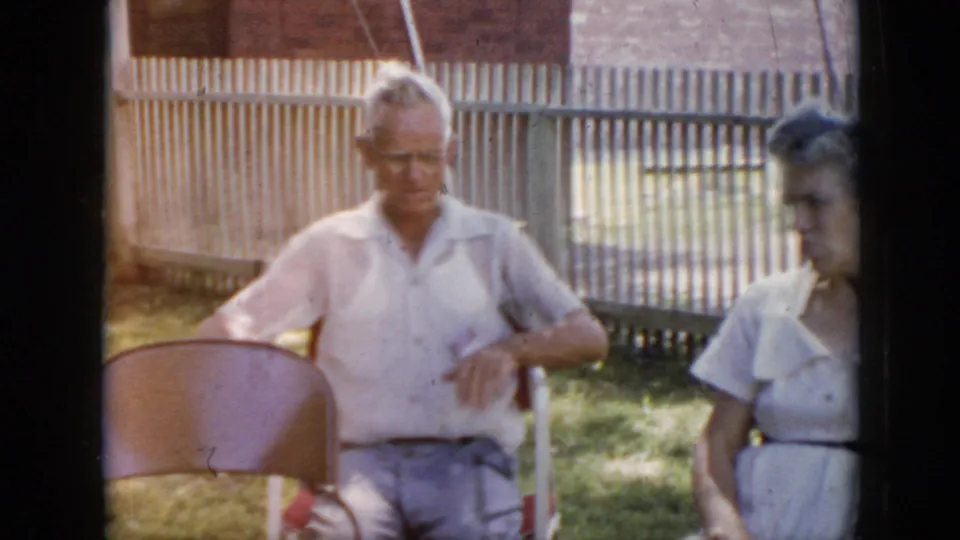 1956 Cuba home-movie frame showing an older man seated near a wooden fence with a woman at the side of the frame.