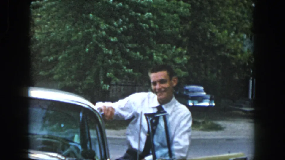 In 1958 Dayton, Ohio home-movie footage, a smiling young man stands by the open side of a yellow vintage car on a residential street.