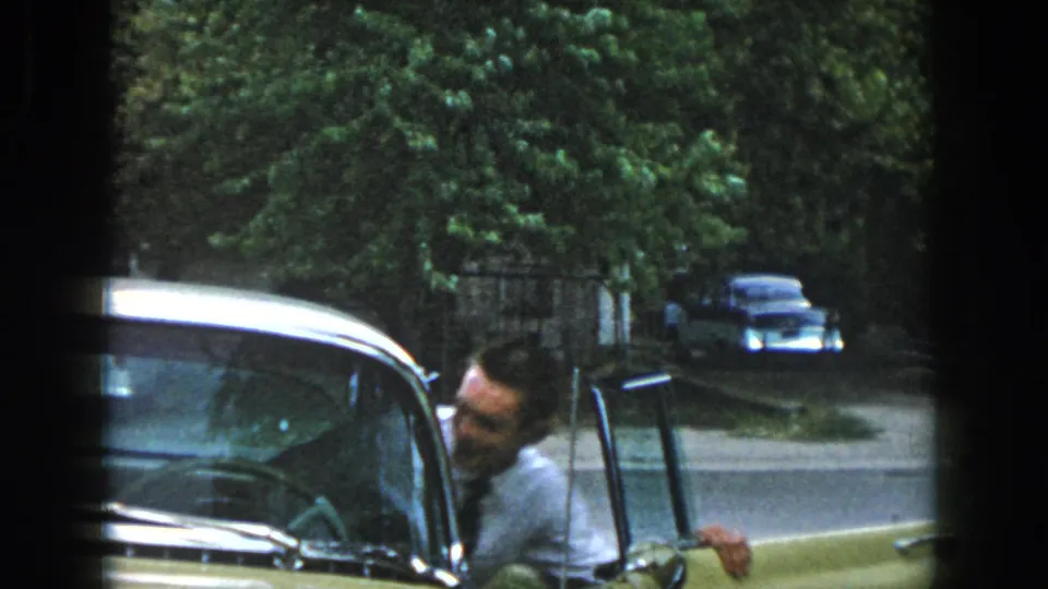 The man leans into or toward the open door of the yellow car, with trees and another older car visible across the street.