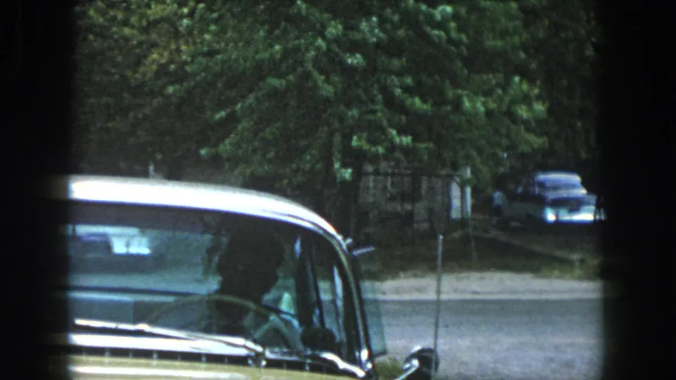 The man is mostly out of view as the yellow vintage car fills the frame, with a quiet street and older car in the background.