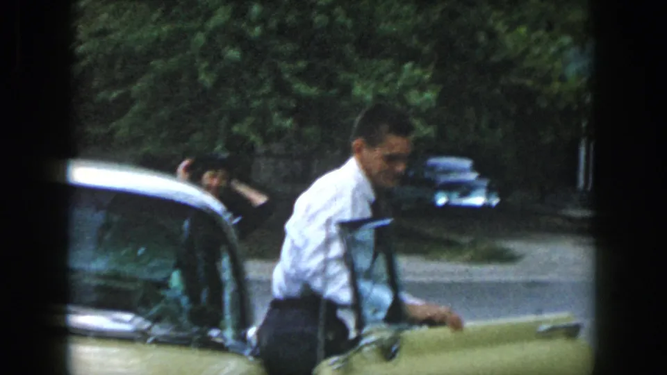 The man stands by the open door of the yellow car in 1958 Dayton footage, with another person faintly visible behind him.