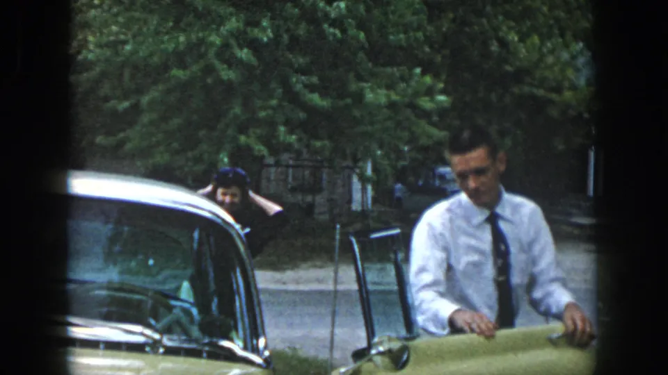 In 1958 Dayton home-movie footage, a young man in a white shirt and tie stands by a light-colored car on a leafy street.
