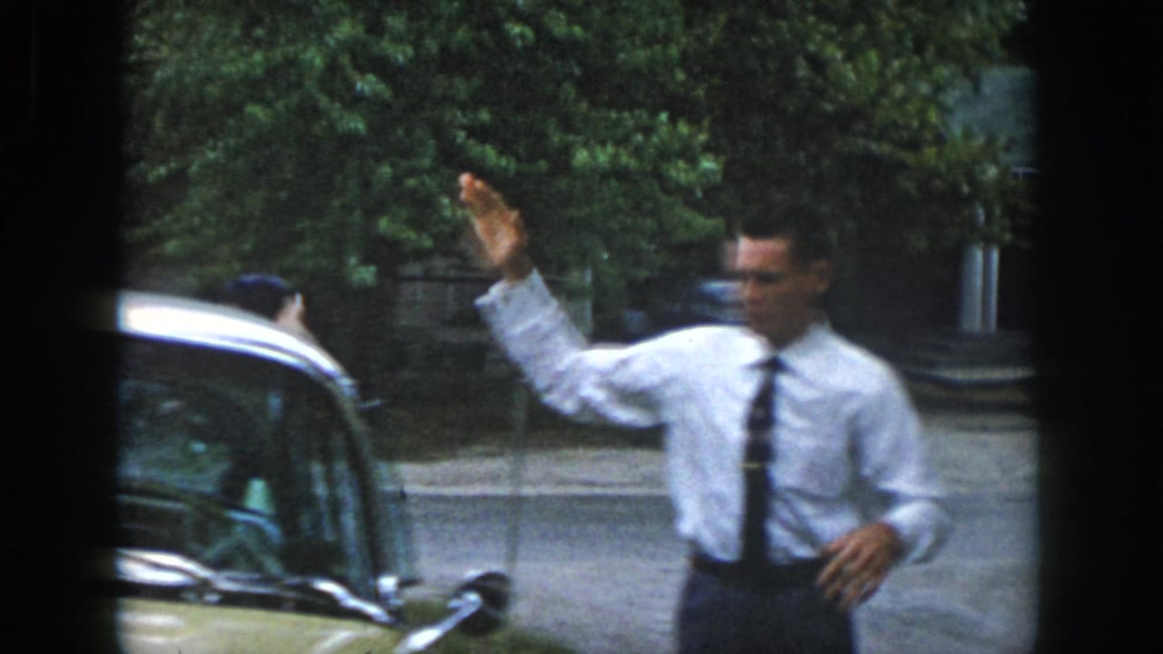 The man raises his hand in greeting beside a mid-century car on a tree-lined Dayton street, with another person partly visible near the vehicle.